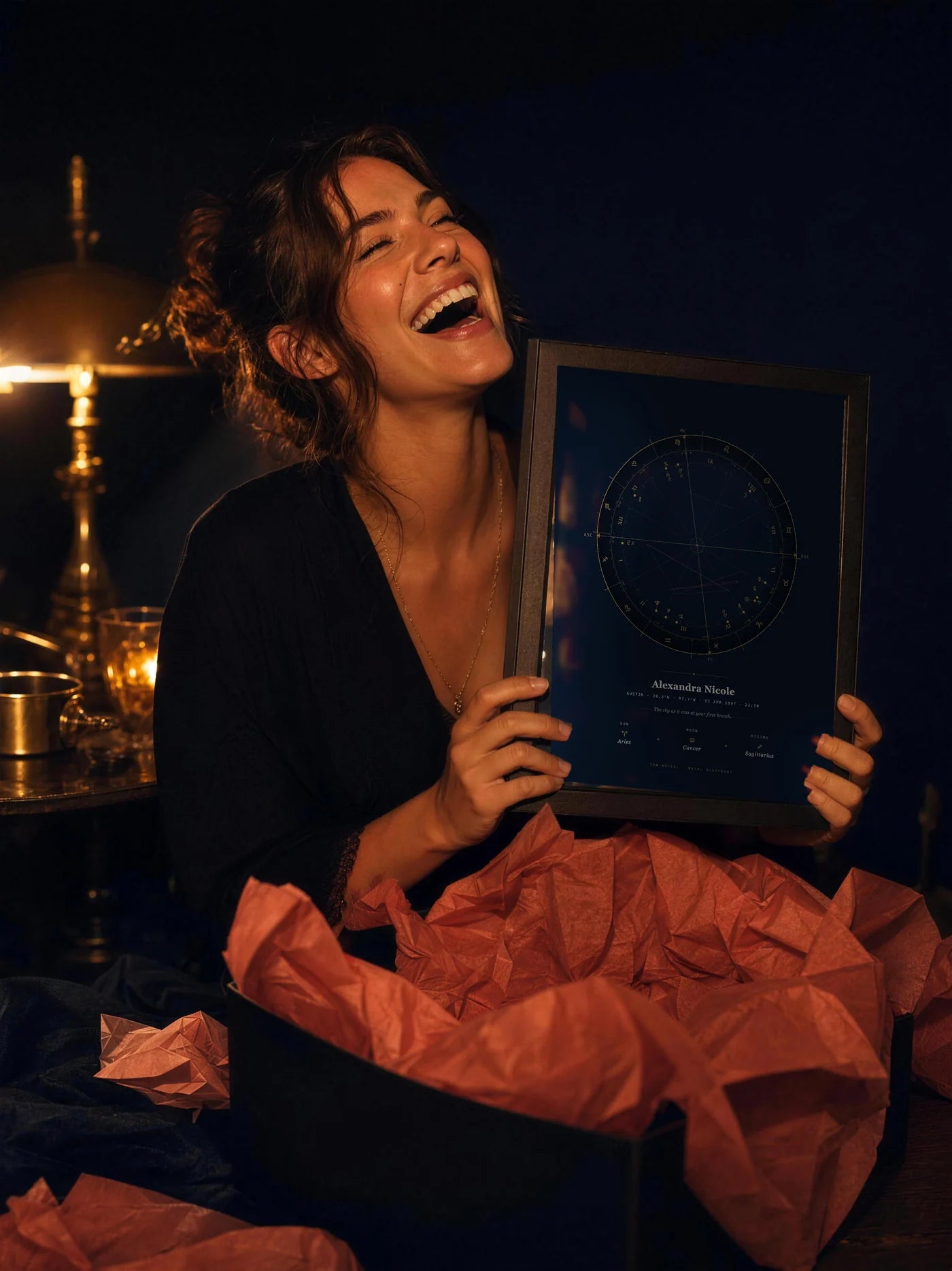 A woman laughs, holding a framed Ion Astral natal blueprint she has just unwrapped, tissue paper around her.