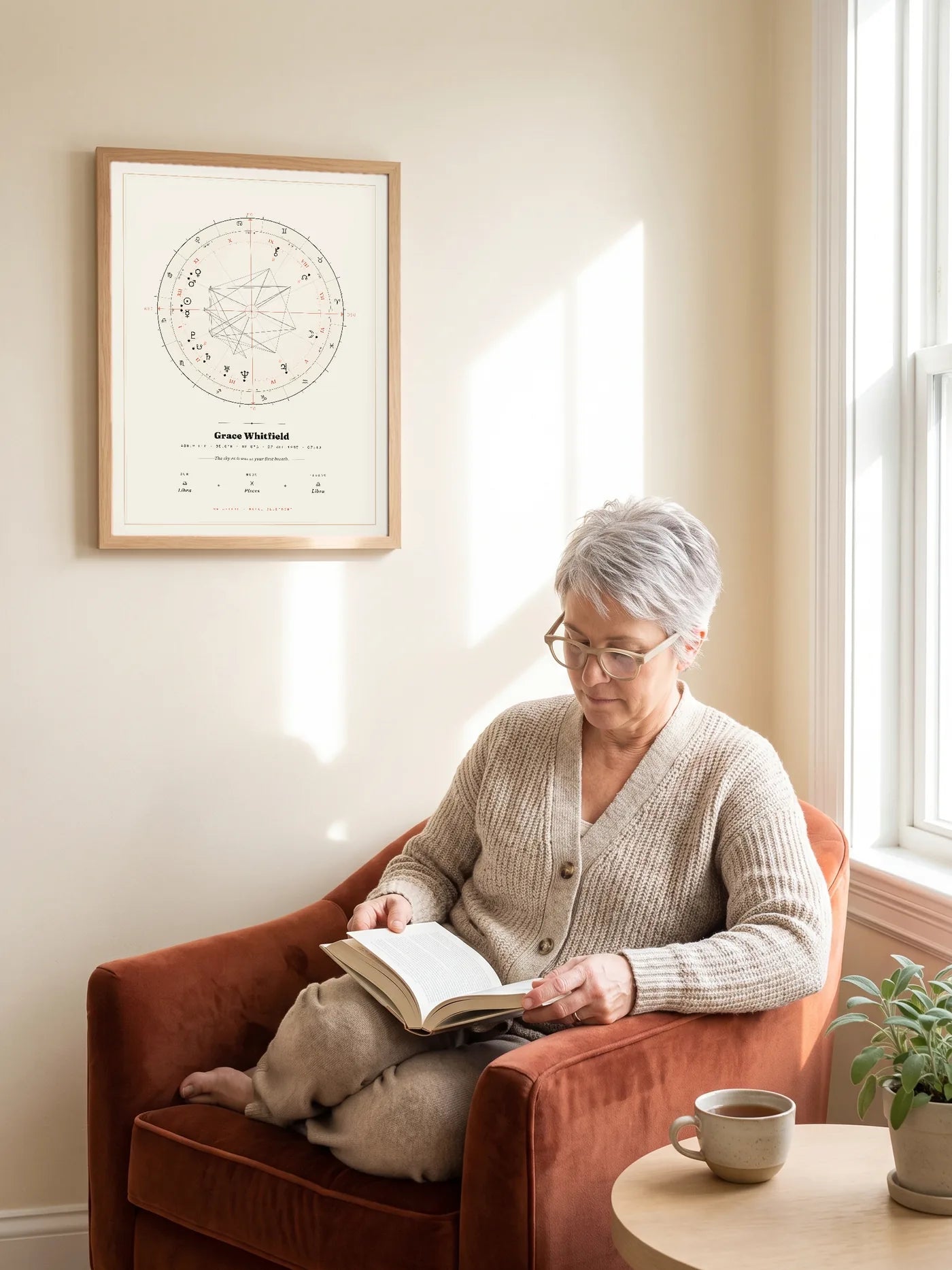 A woman with silver hair reads in a rust armchair below an oak-tone framed Natal Blueprint in the cream Atelier style, morning sun on the wall.