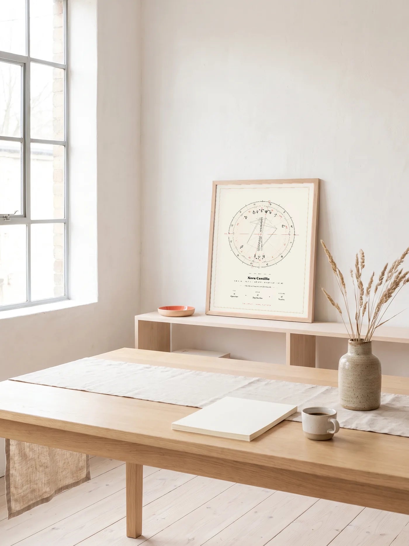 An oak-tone framed Natal Blueprint in the Atelier style leans on a pale shelf in a sunlit studio, behind a wooden worktable.