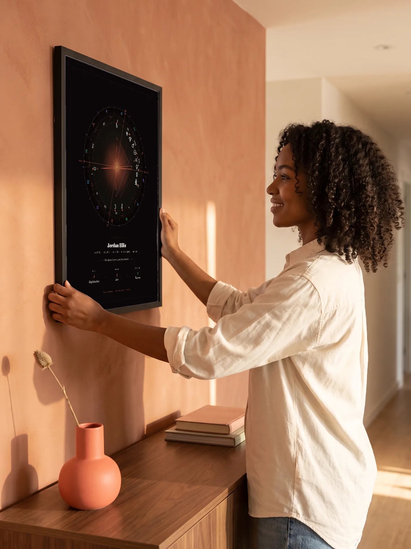 A woman hangs a black-framed Natal Blueprint in the Element style, near-black with a coral glow, on a terracotta wall.