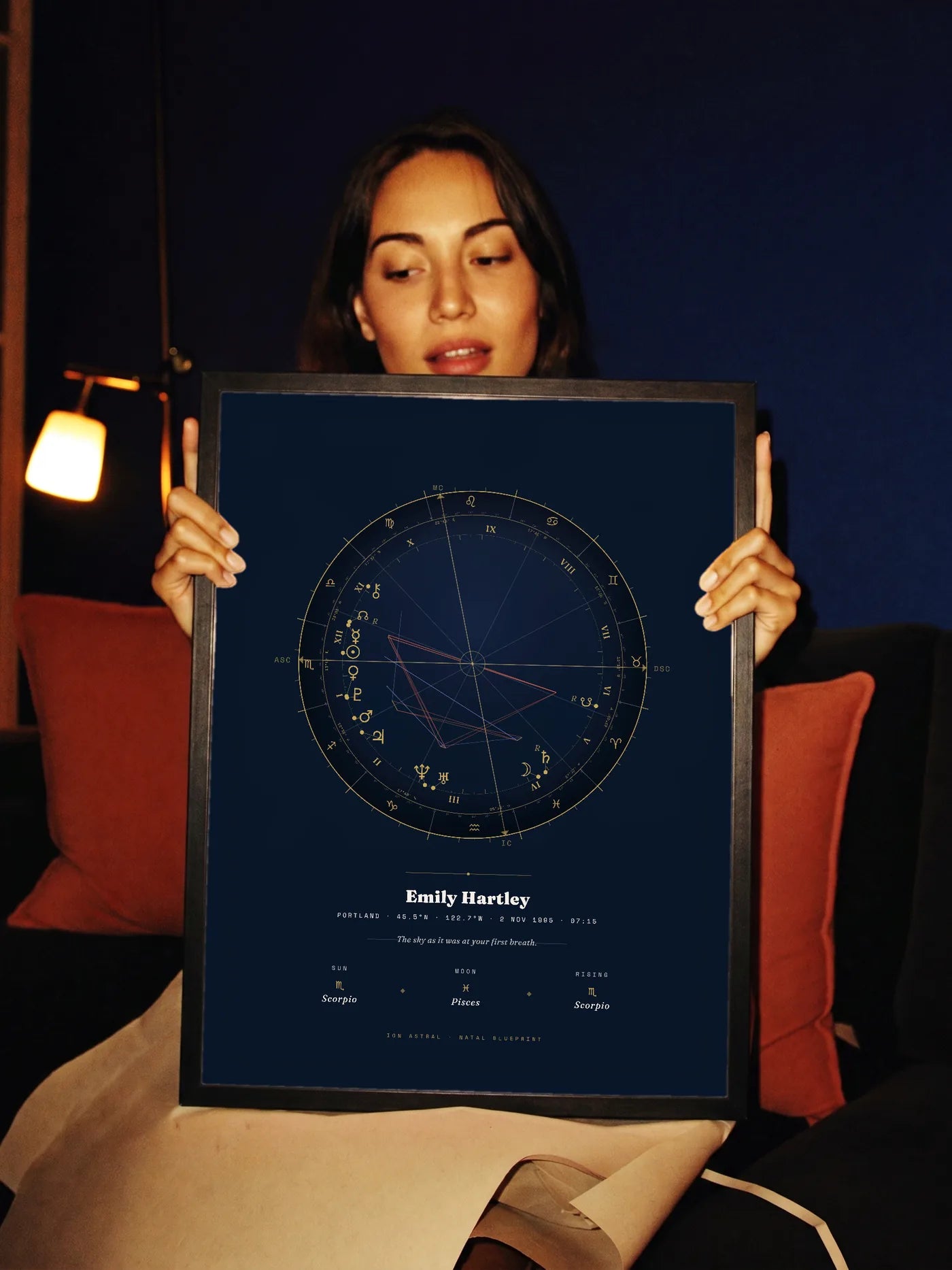 A woman in lamplight holds up a black-framed Natal Blueprint in the Midnight Gold style, navy with gold linework.
