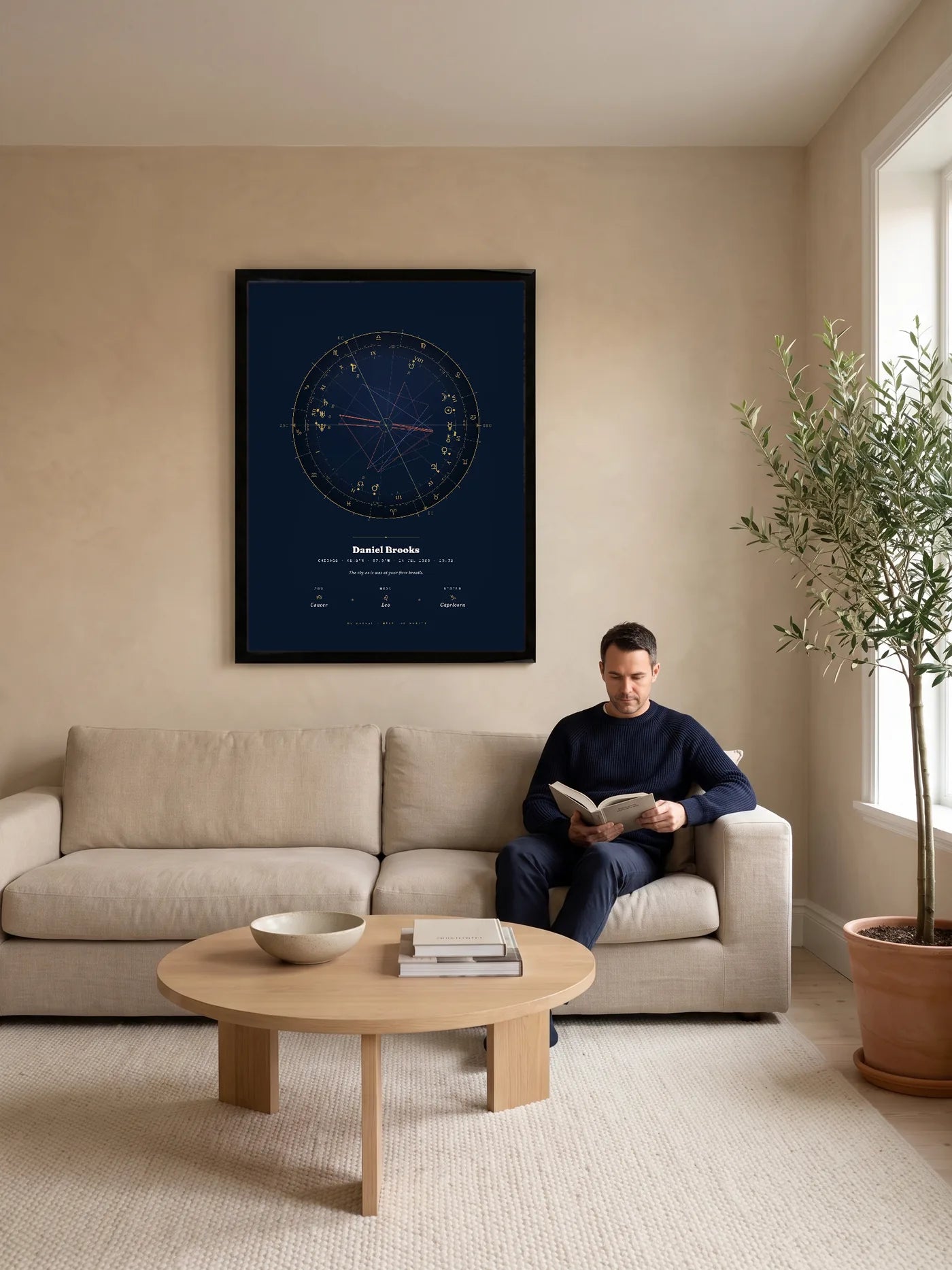 A bright living room with a large black-framed Natal Blueprint in the Midnight Gold style above a linen sofa, where a man sits reading.