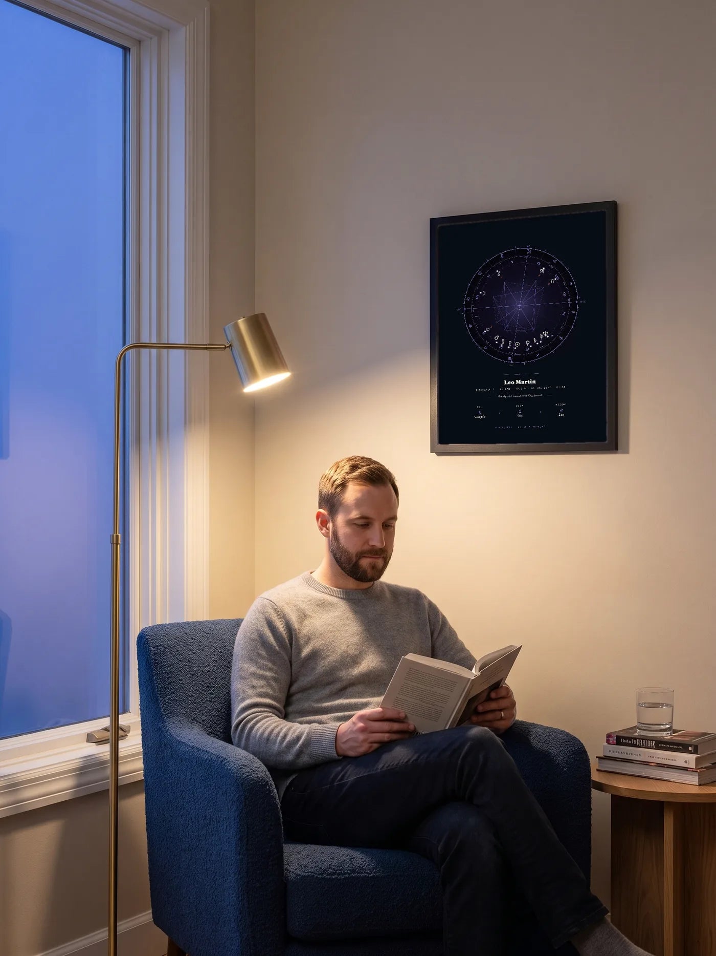 At dusk, a man reads in a blue armchair beneath a lamp-lit, black-framed Natal Blueprint in the Nocturne style.